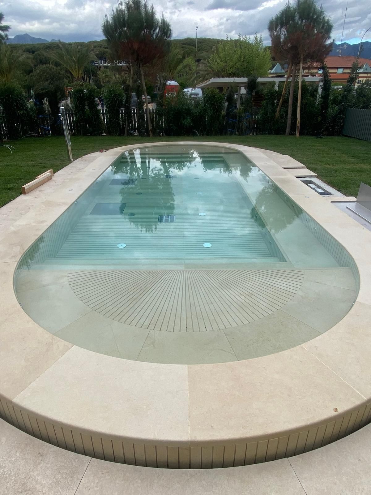 Piscina di forma ovale con bordo in pietra, parzialmente coperta da una copertura retrattile. Prato verde e alberi sullo sfondo.
