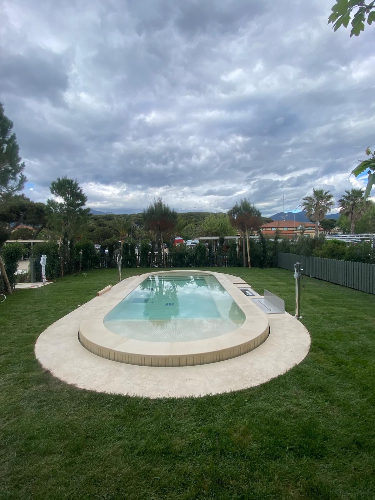 Piscina ovale in un giardino erboso, circondata da cemento color sabbia. Cielo nuvoloso in alto.