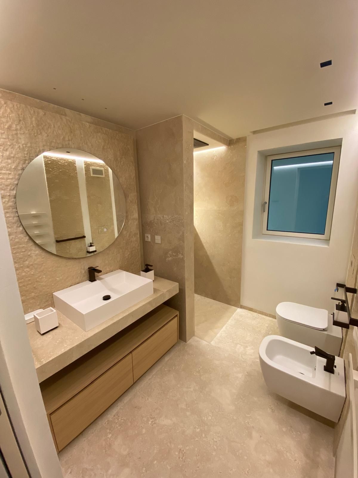 Bagno moderno con specchio rotondo sopra il lavandino, il WC e il bidet. Tonalità beige e bianco, sanitari neri.