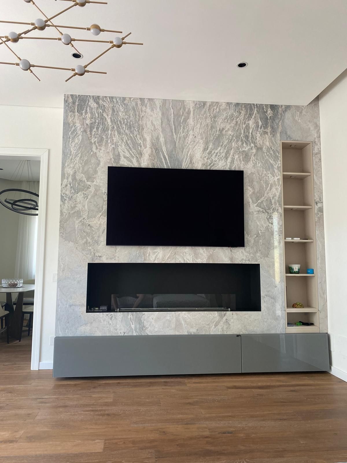 Parete in marmo grigio con TV nera, camino, mensole a muro. Pavimento in legno scuro, mobili grigi. Interni moderni.