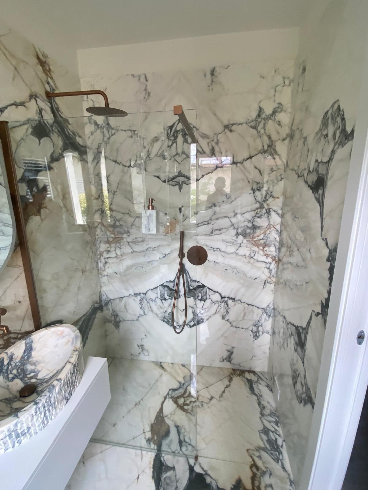 Doccia rivestita in marmo con porta in vetro trasparente e rubinetteria in bronzo. Lavandino con piano in marmo visibile a sinistra.