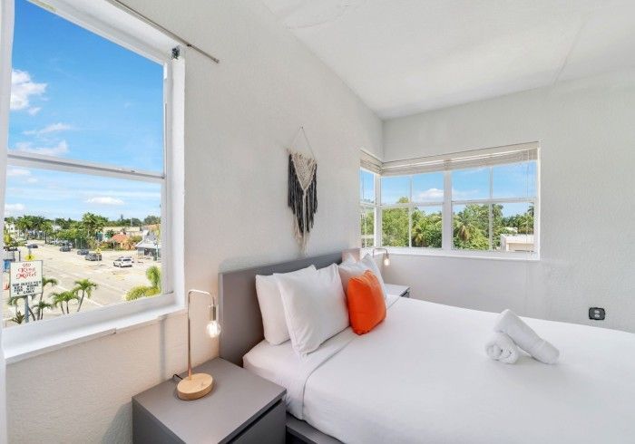 Bedroom with bed, pillows, and windows overlooking a street scene. White walls, grey furniture, orange pillow.