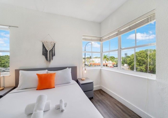Bedroom with a bed, two windows with a view, and neutral colors with an orange pillow.