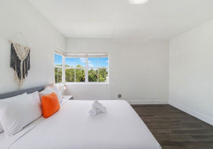 Bedroom with white walls, a bed with white and orange pillows, a macrame wall hanging, and a window with a view.