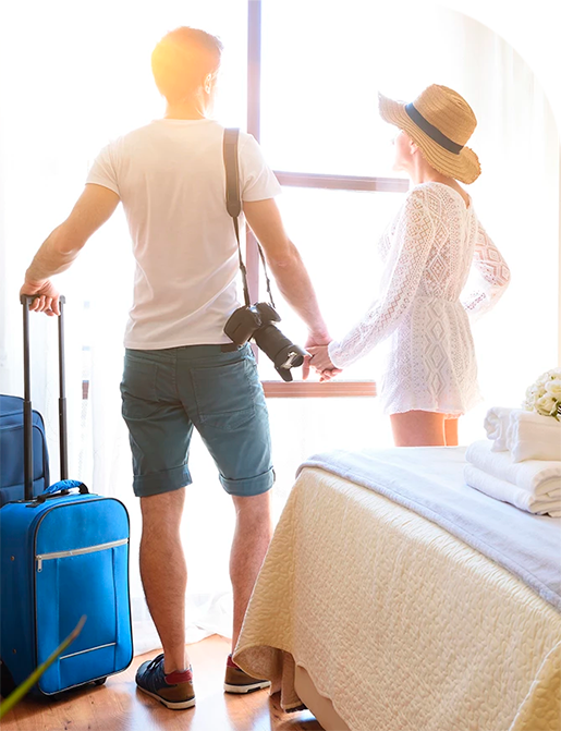 Couple holding hands, looking out a window. Man holds luggage, camera. Woman wears a sun hat and white dress.