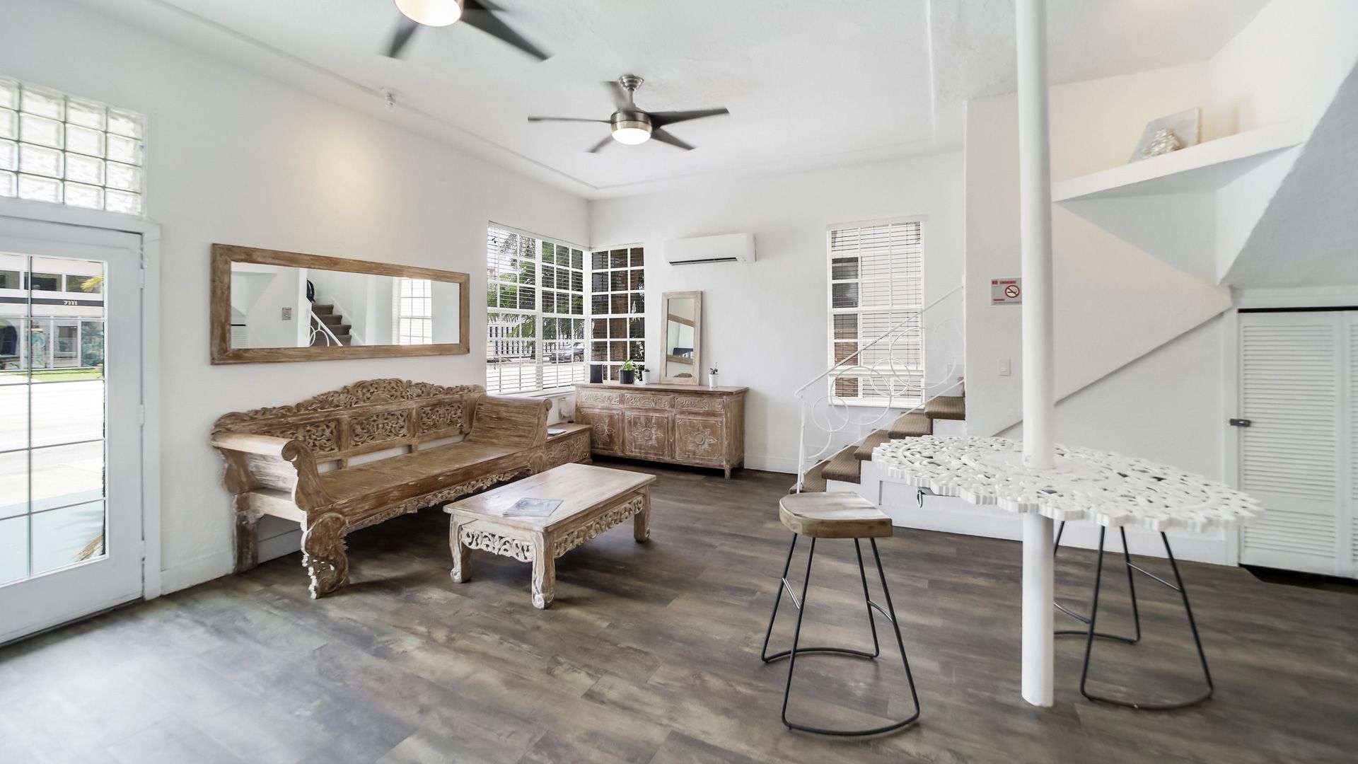 Bright, open interior with white walls, wood floors, and carved wood furniture. A staircase leads to a loft area.