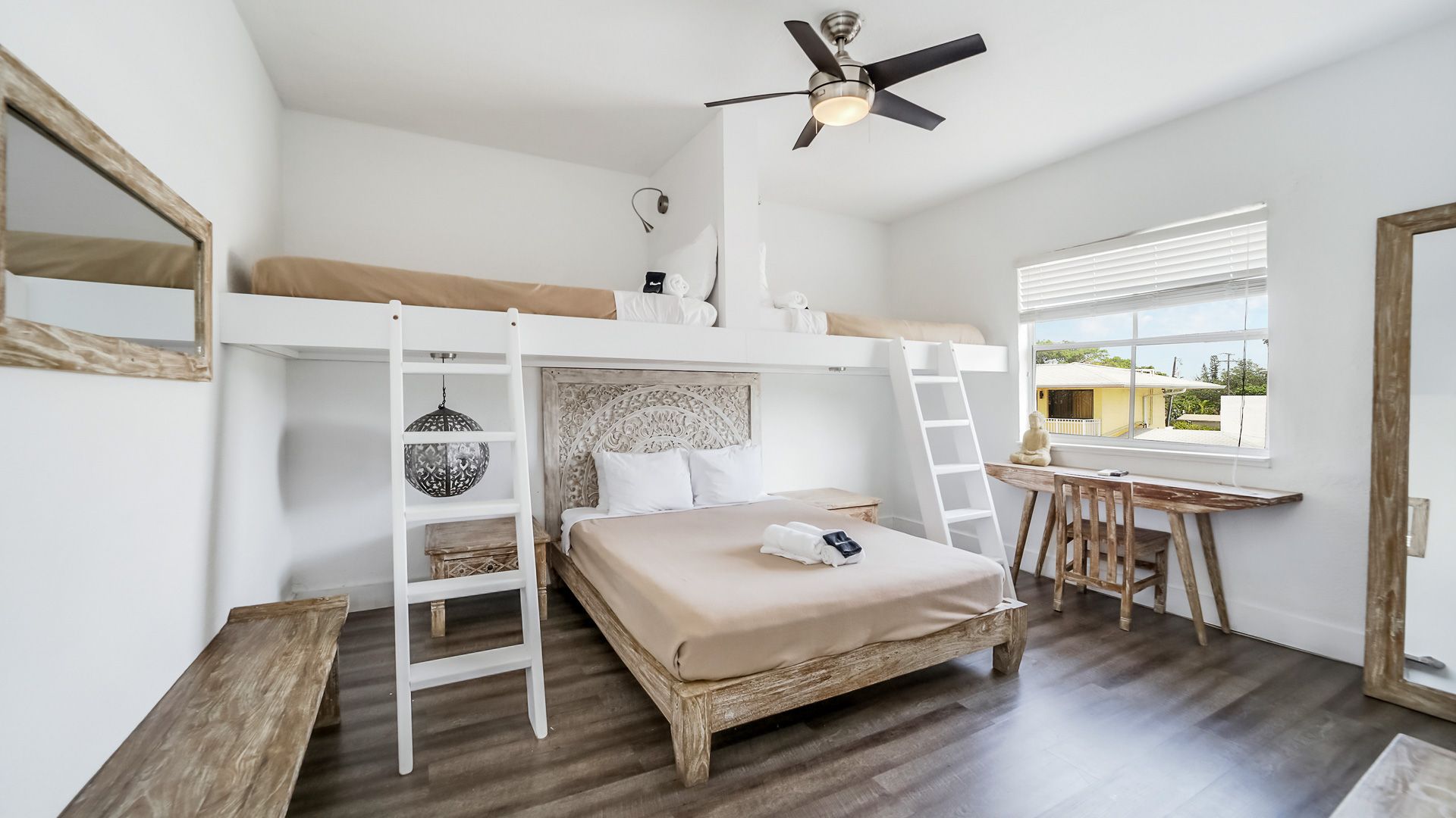 Bright bedroom with loft bed, desk, and large window. Wooden accents, white walls, and dark wood floor.