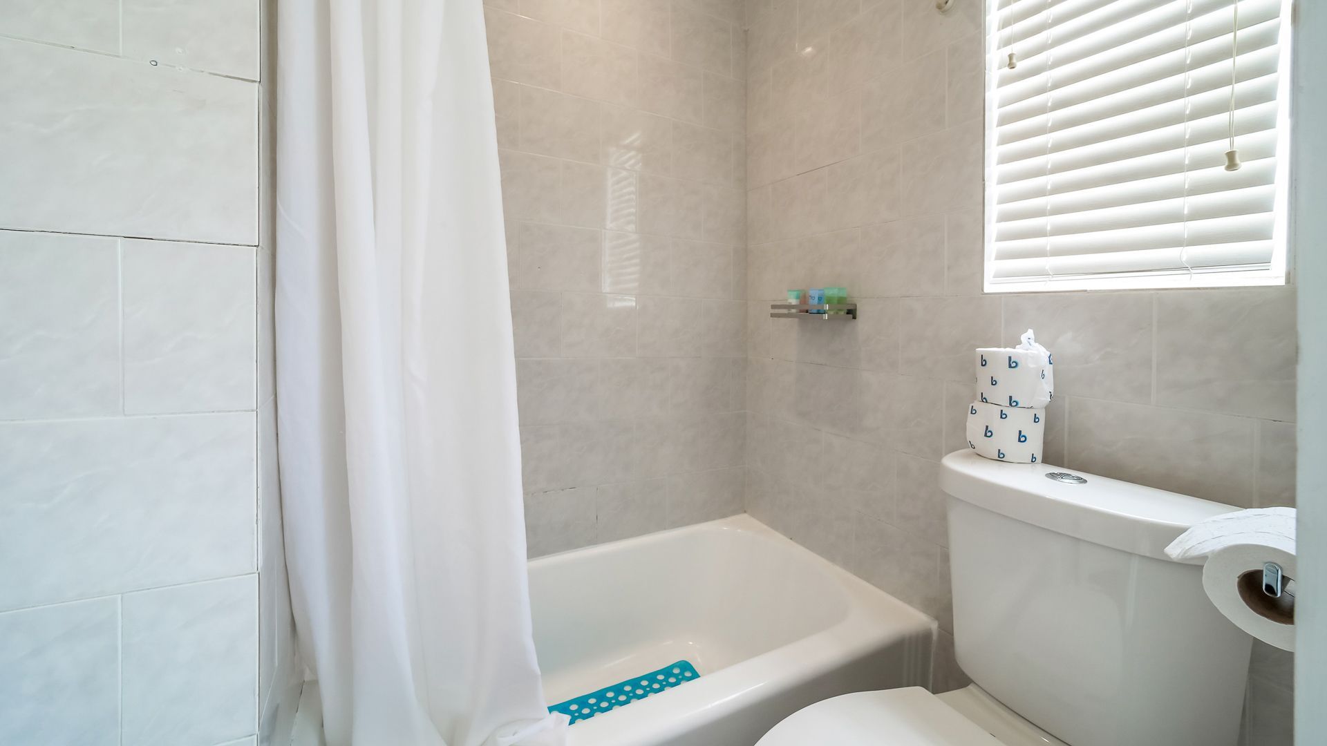 Bathroom with bathtub, toilet, and window with blinds. White walls and shower curtain.