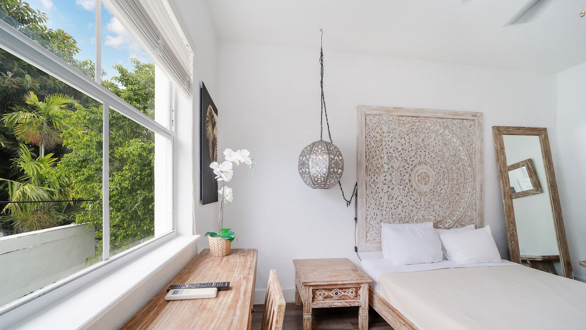 Bedroom with a bed, desk, and large window overlooking trees. White walls, wood furniture, light tones.