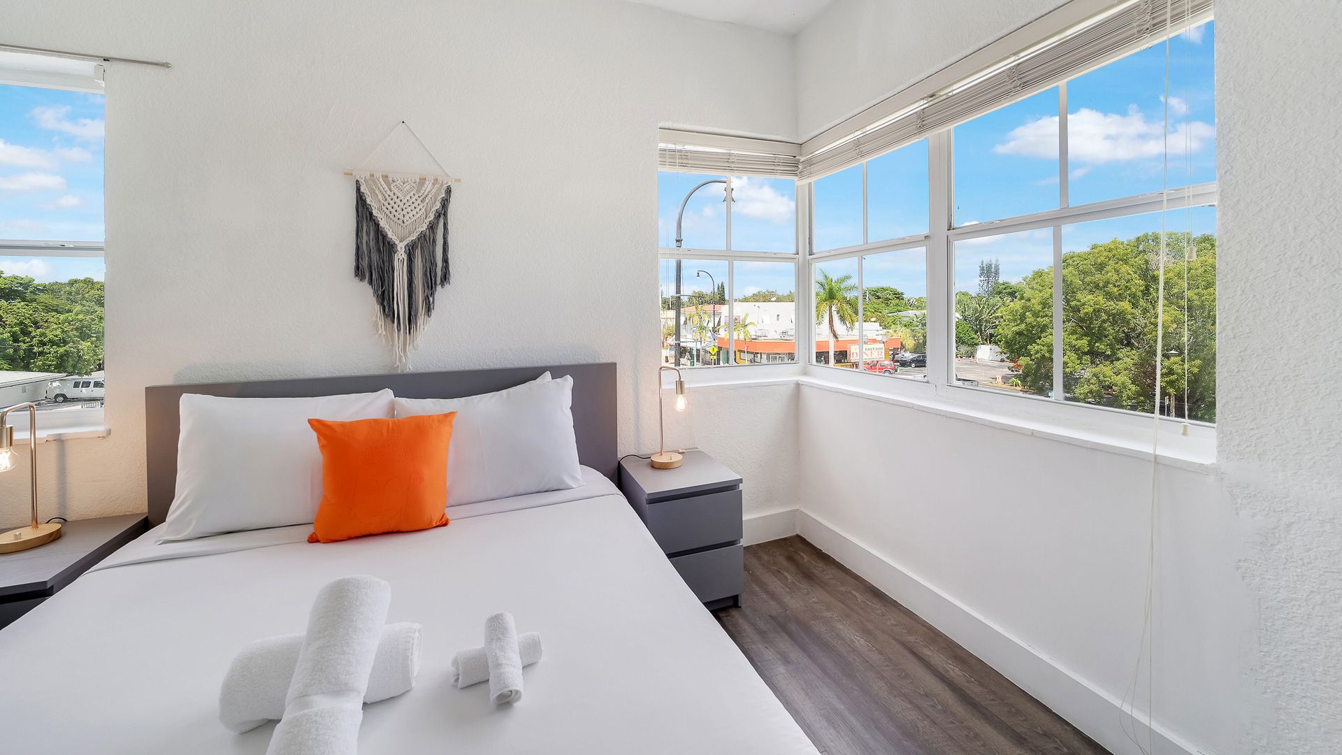 Bedroom with white walls, a bed with an orange pillow, and large windows overlooking a city scene.