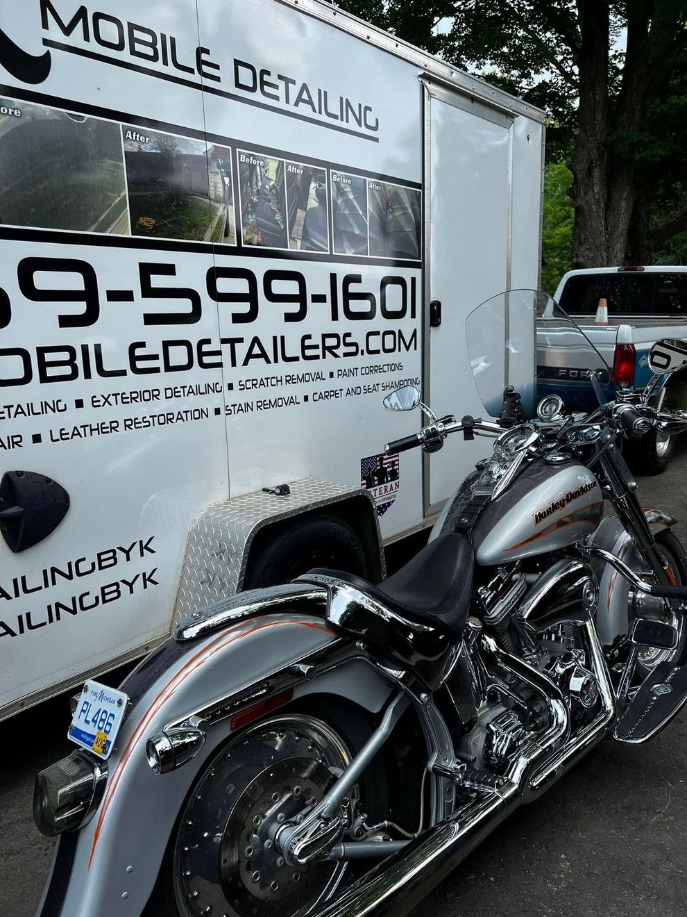 A shiny silver motorcycle parked next to a white mobile detailing trailer with contact information on its side.