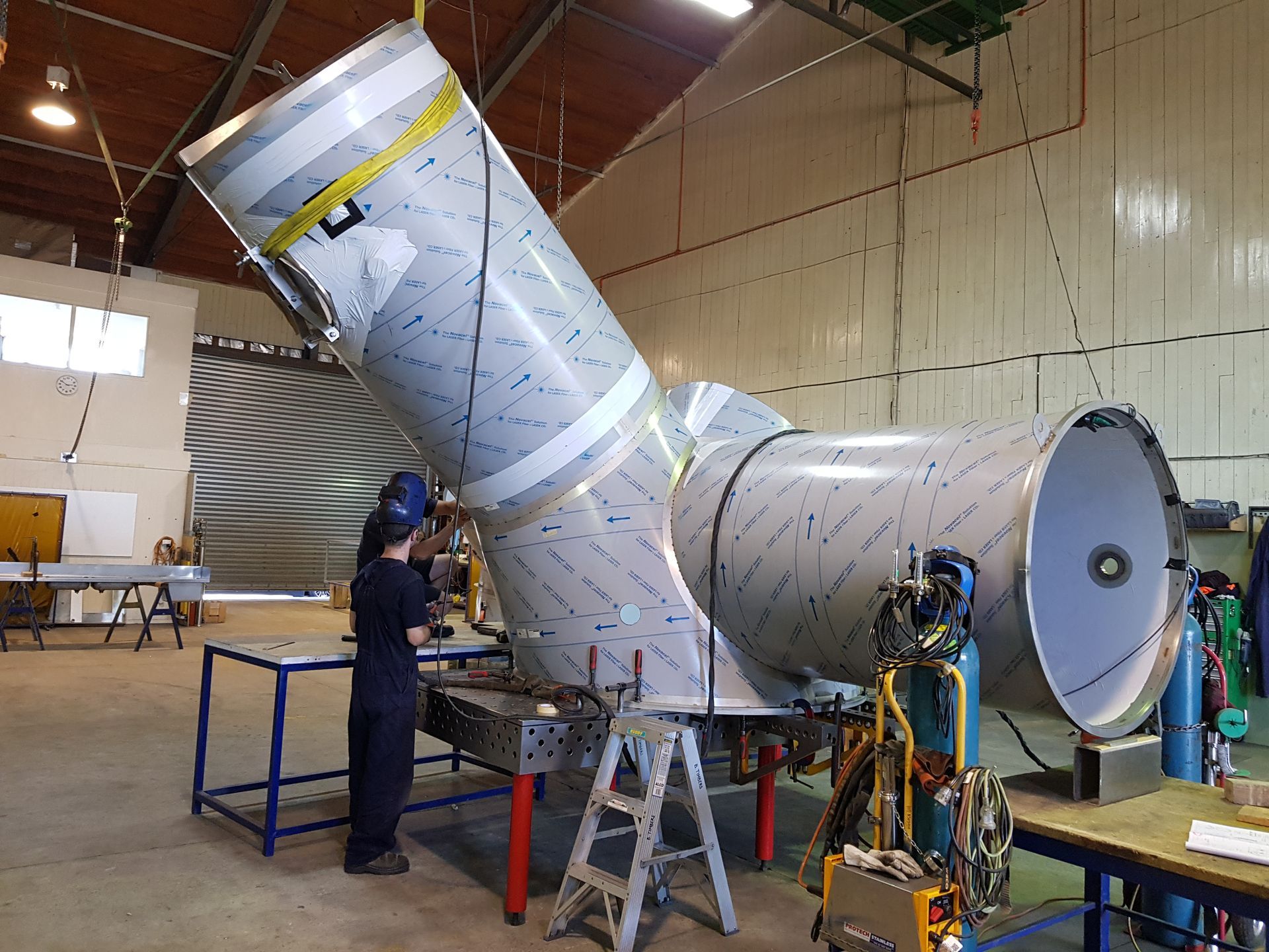 Person welding a large, angled metal structure in a workshop, with tanks and tools visible.