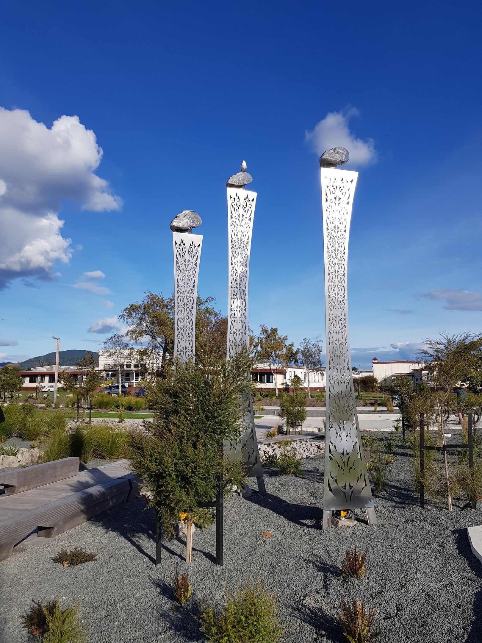 Three tall, silver, patterned sculptures with rocks on top, set in a gravel garden under a blue sky.
