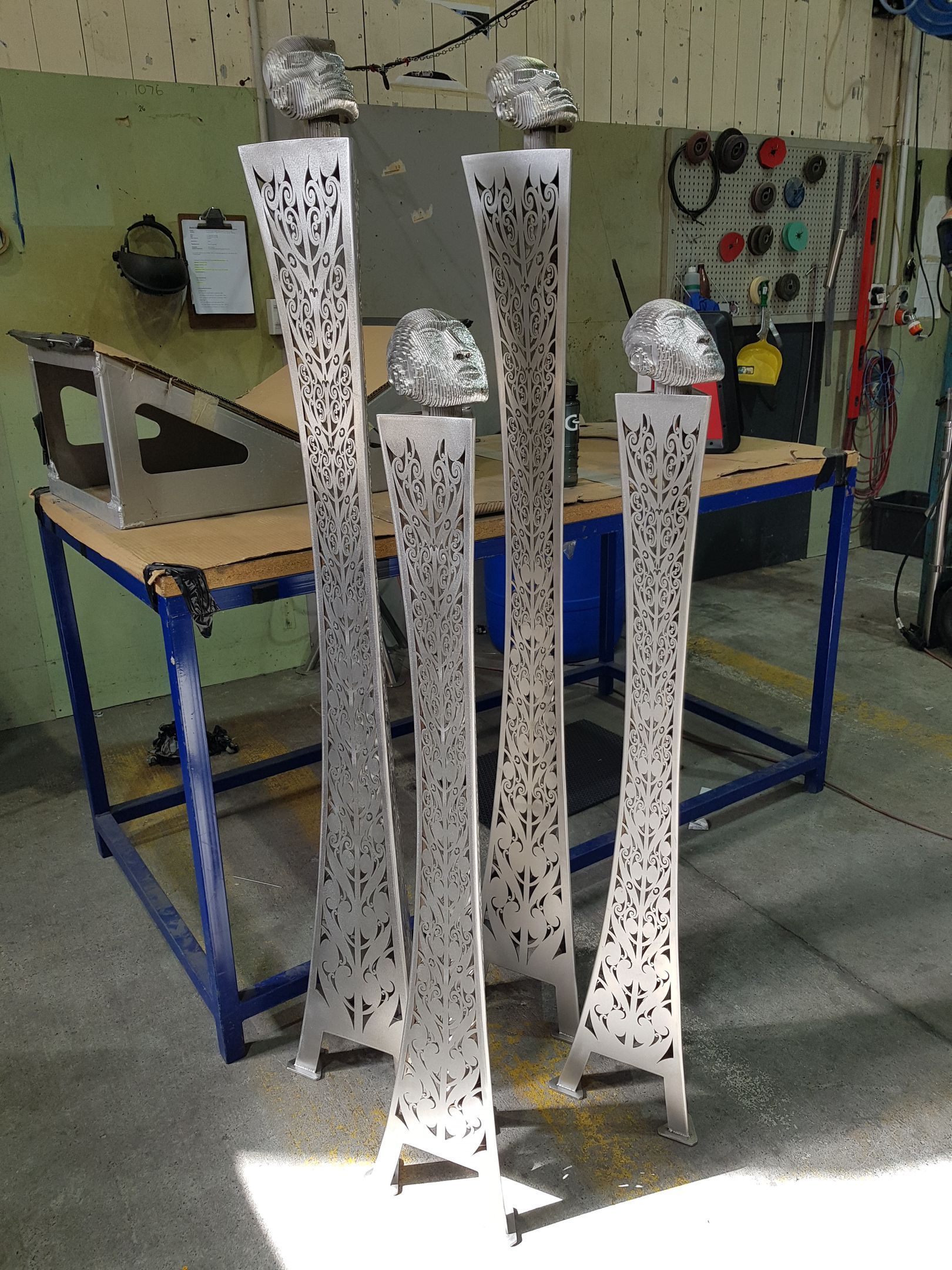 Five tall, metal sculptures with patterned cutouts in a workshop. Each has a spherical top.