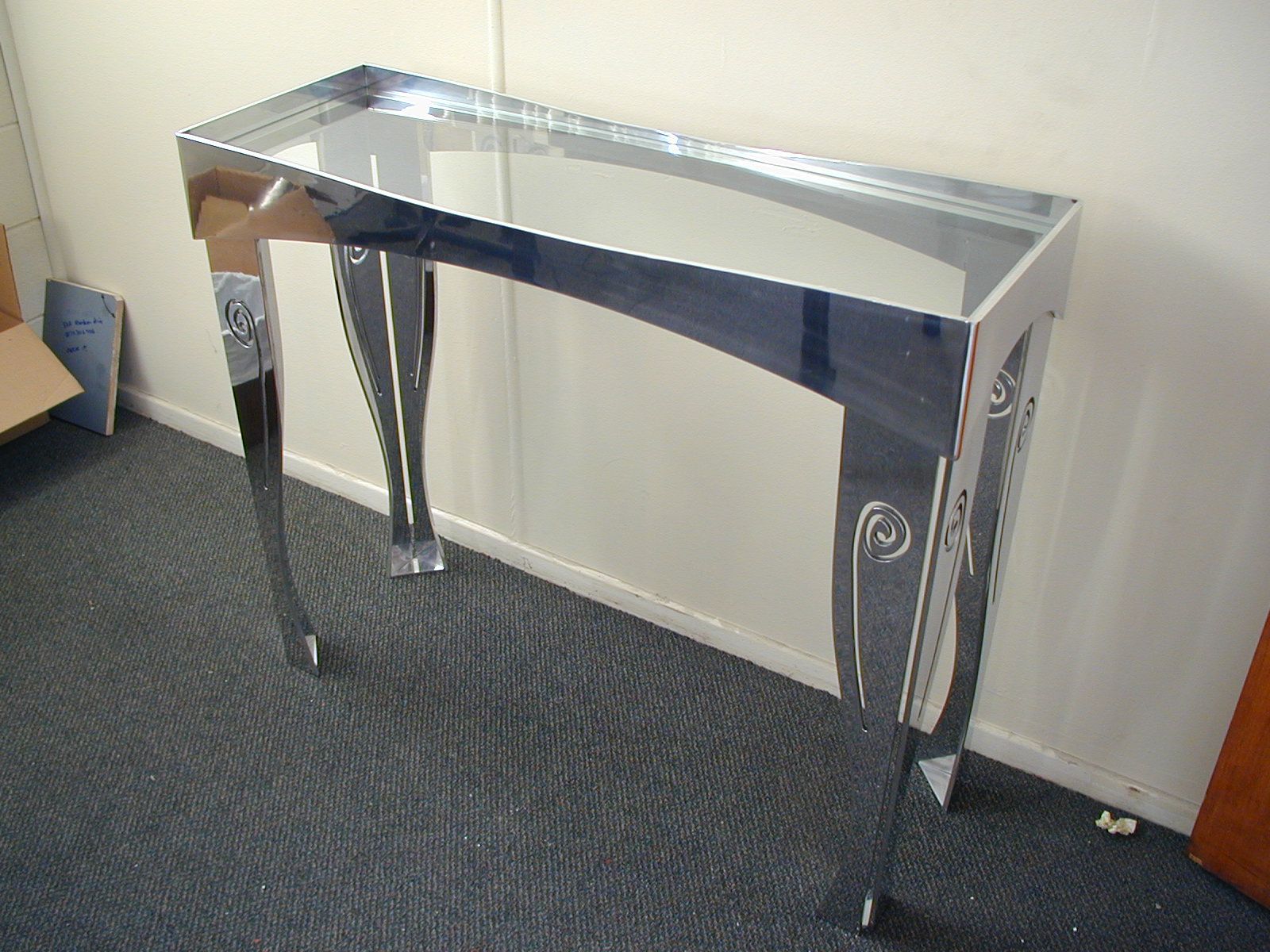 Shiny silver console table with ornate legs, set against a white wall on a dark carpet.