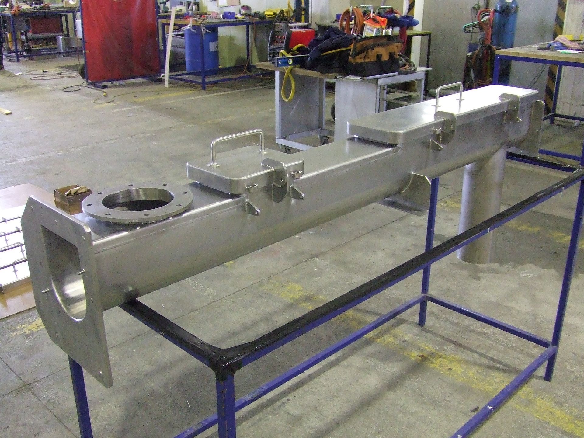 A large metal fabricated component, likely industrial, sits on a blue metal stand in a workshop.