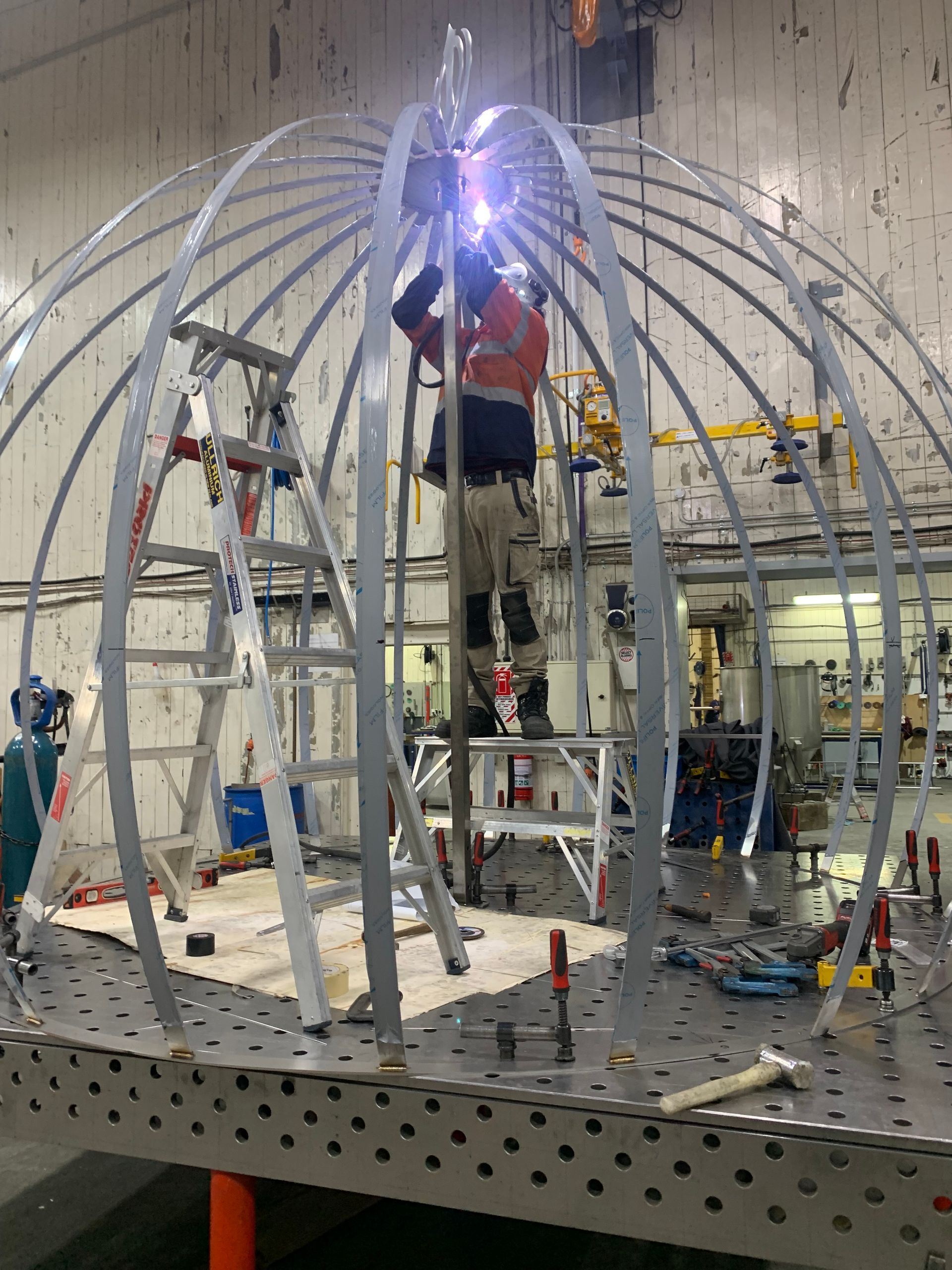 A person welding metal dome frame, standing on a ladder in a workshop.