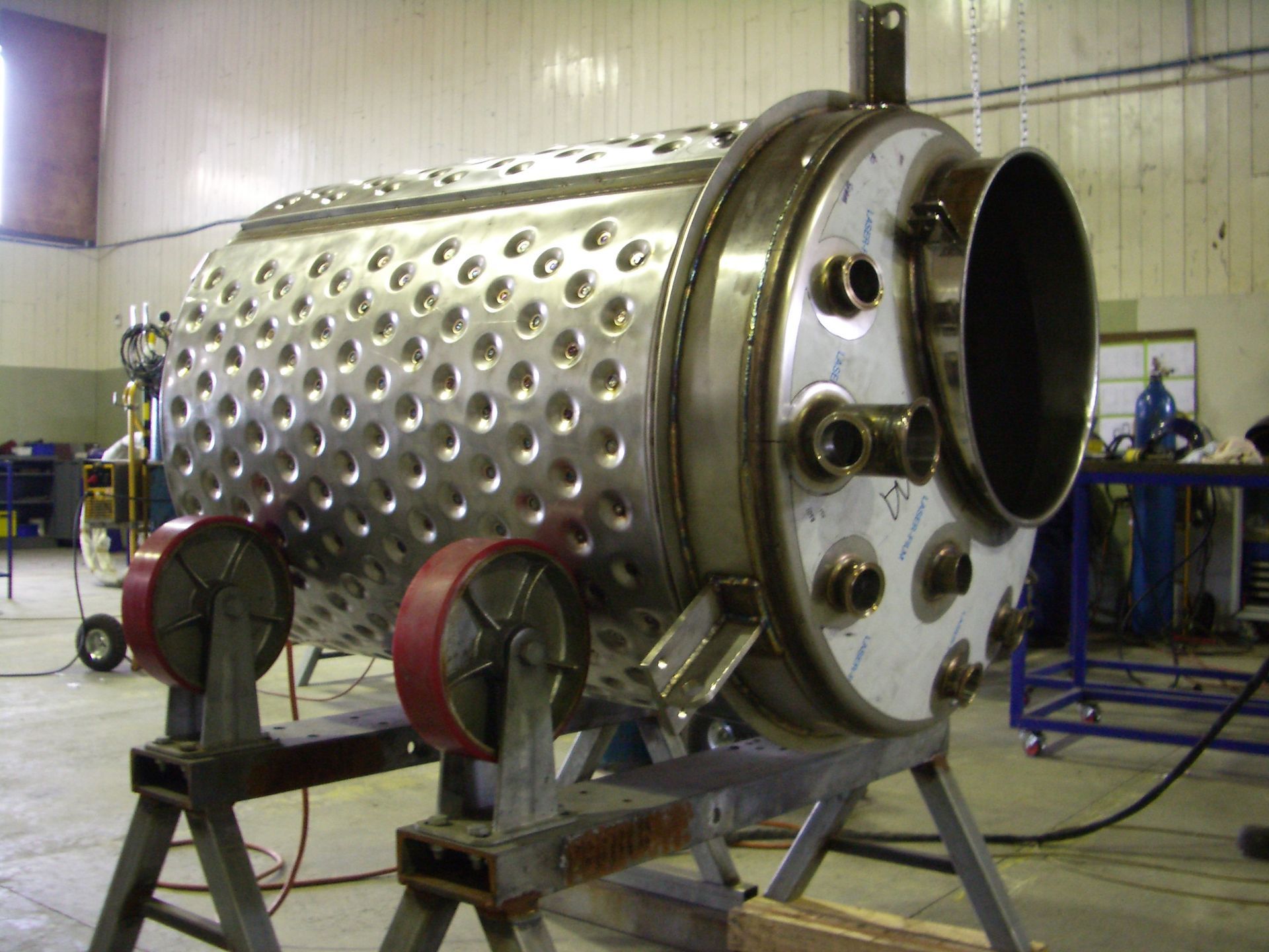 Large cylindrical metal tank with numerous holes and side attachments, mounted on rolling supports in a workshop.