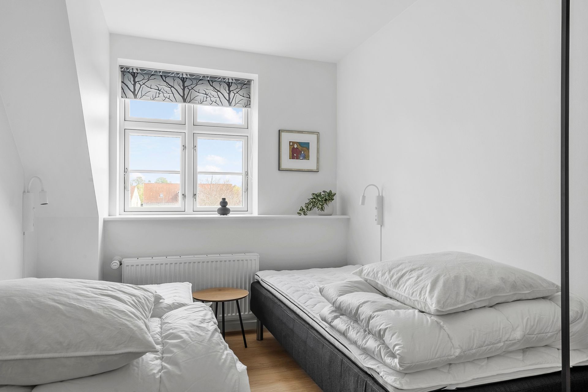 Soveværelse med to senge, lille bord, vindue med mønstret skygge, hvide vægge.