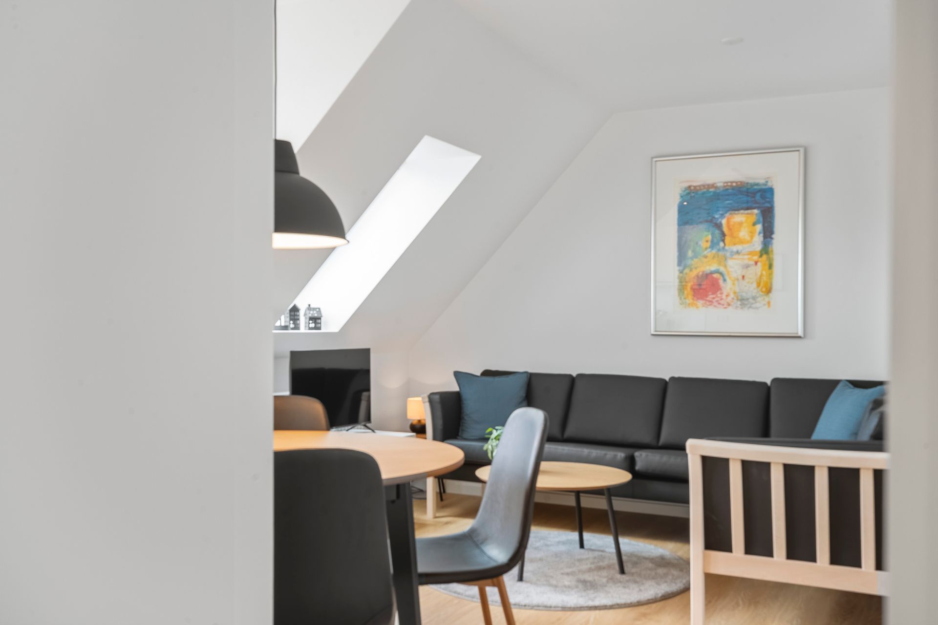 Stue med sort sofa, rundt bord, kunst og ovenlysvindue.