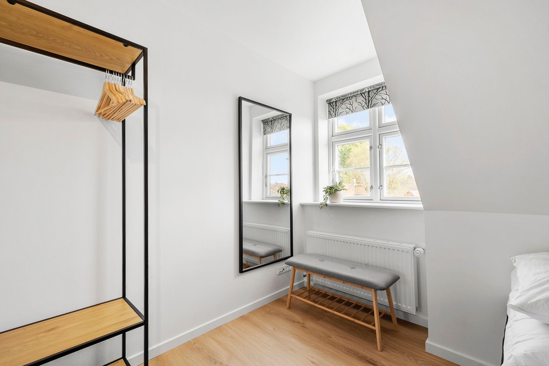 Et minimalistisk soveværelse med et spejl i fuld længde, bænk, åbent skab og vindue med naturligt lys.