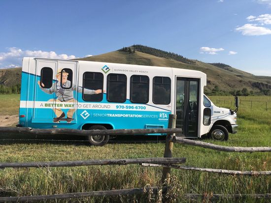 White and teal passenger bus in a field, door open, with a mountain in the background.