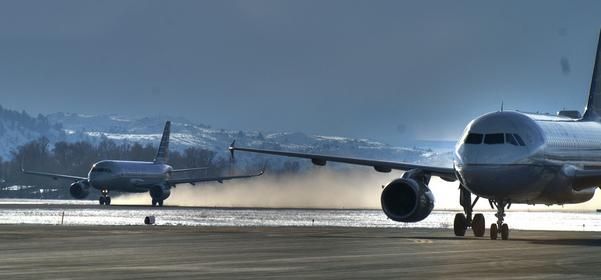 Two airplanes on a runway, likely taxiing. The sky is blue and a snowy hill can be seen in the distance.