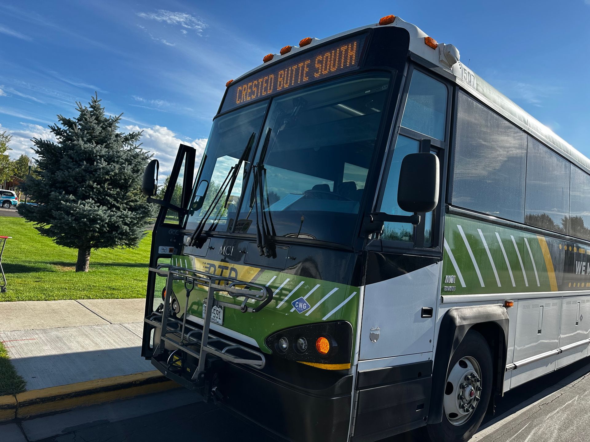 Bus parked, door open. Green and white exterior, digital route display above windshield.