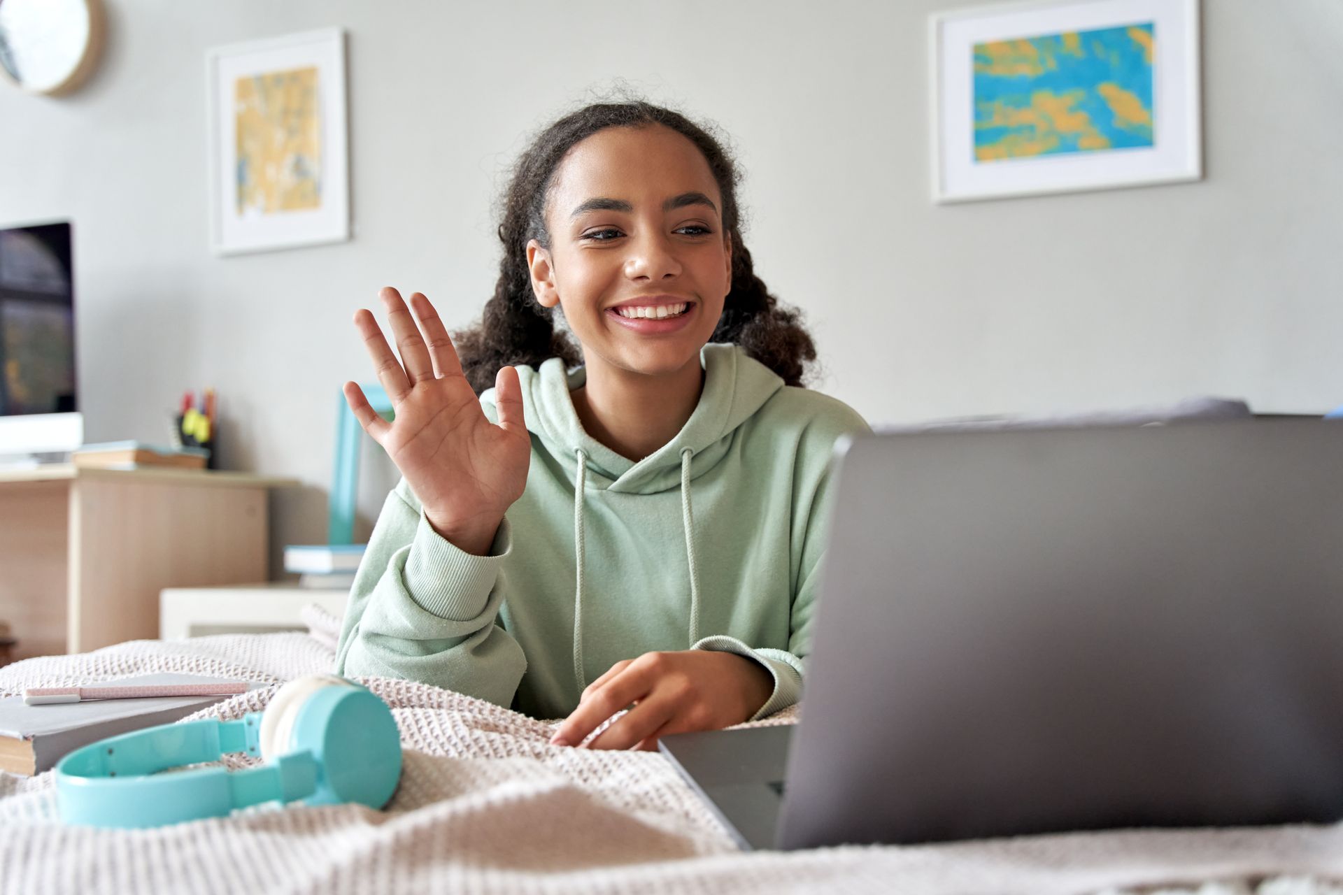 Young person waving on a video call, wearing a light green hoodie, indoors.