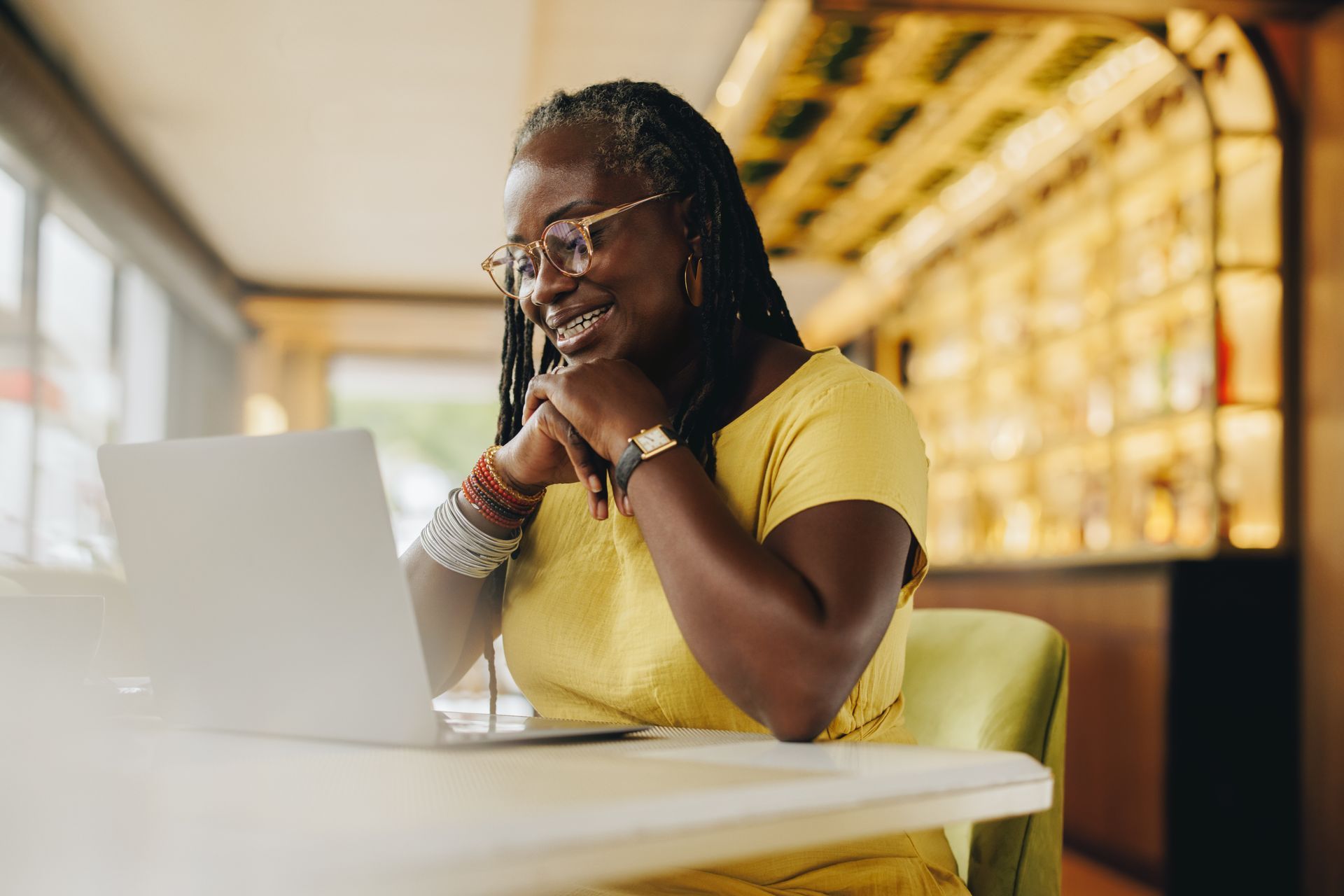 Woman smiling while looking at a laptop, seated at a table in a cafe.