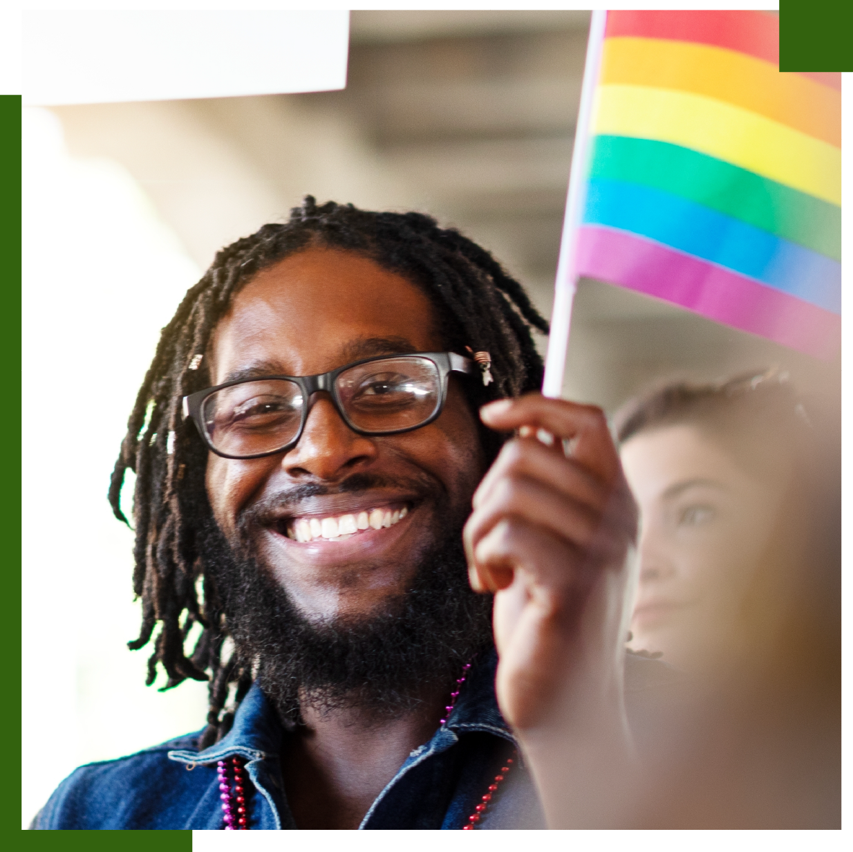 Man with dreadlocks and glasses smiles, holding a rainbow flag.