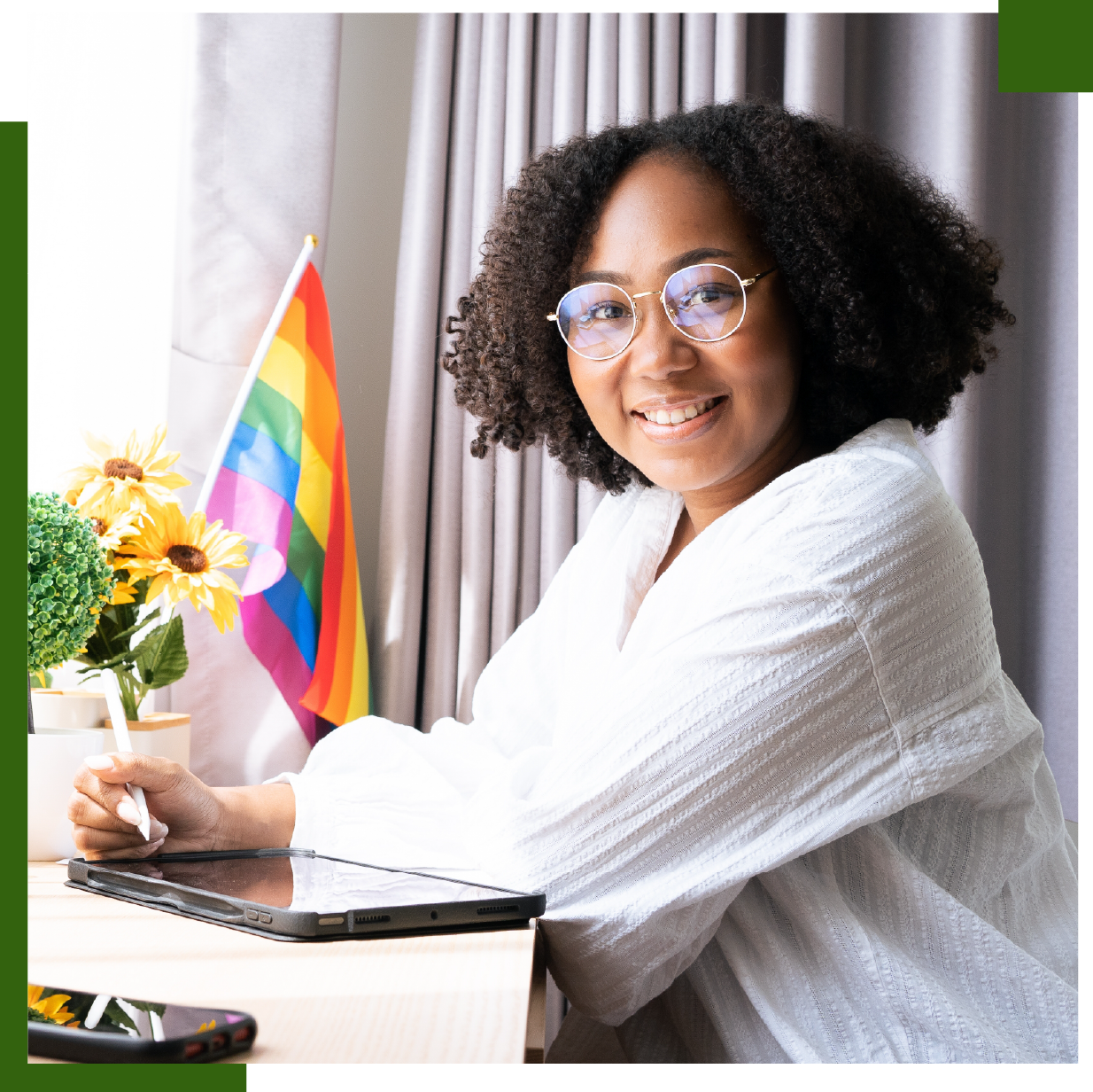 Person with glasses smiles while using a tablet, rainbow flag in background.