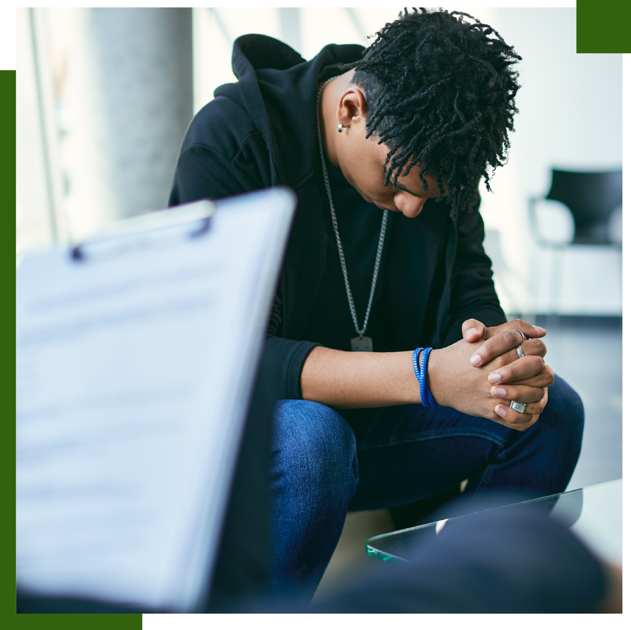Person in a black hoodie and jeans, hands clasped, distressed in a therapy session.