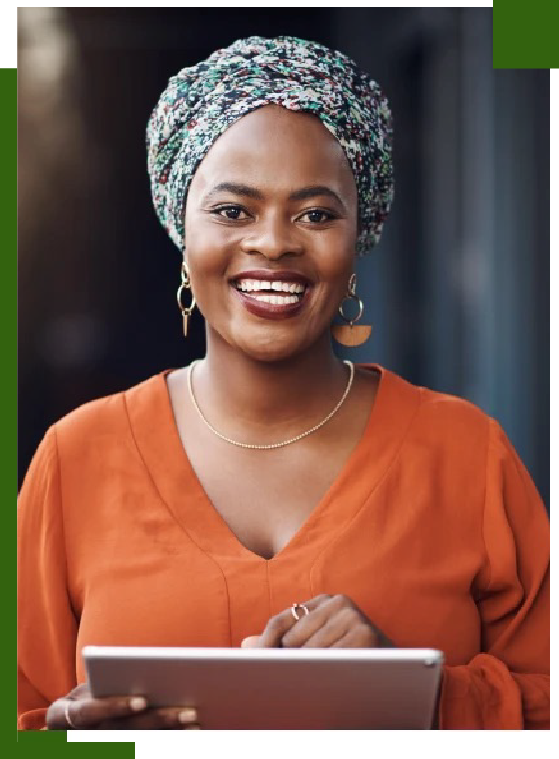 Woman wearing a patterned head covering and orange shirt, holding a tablet and smiling.