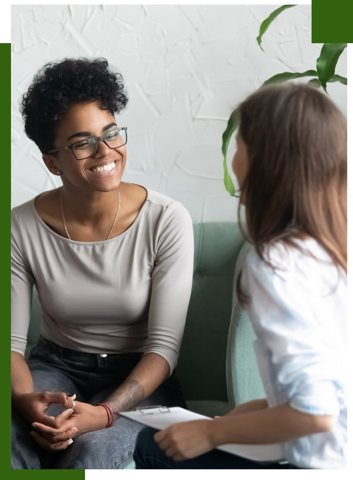Woman smiling during a counseling session. She is sitting on a sofa and looking at the counselor, who holds a clipboard.
