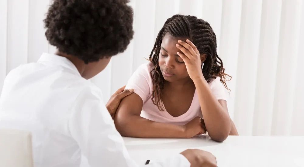 Person comforts another with hand on shoulder; indoors at a white table. Both are looking down.