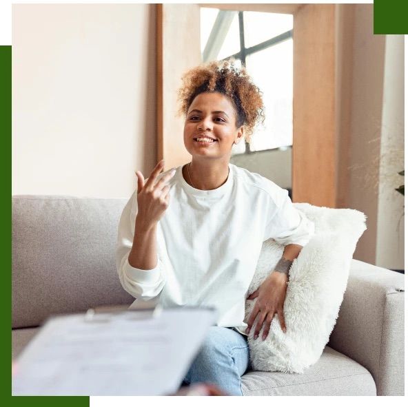 Woman gestures while sitting on a couch, holding a clipboard in a well-lit room.