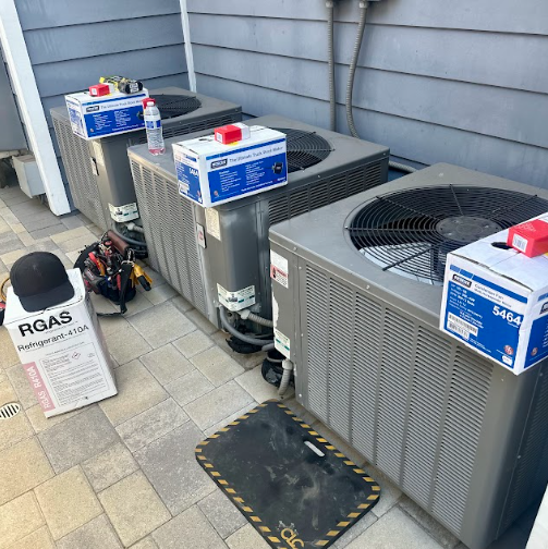 Three air conditioning units outside, with tools and boxes on the ground, likely being serviced.