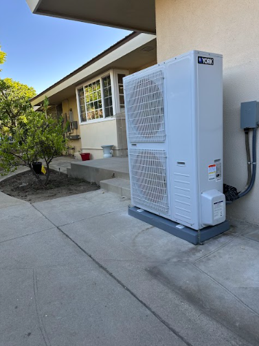 A white HVAC unit on a gray base, mounted on a wall near a house's entrance.
