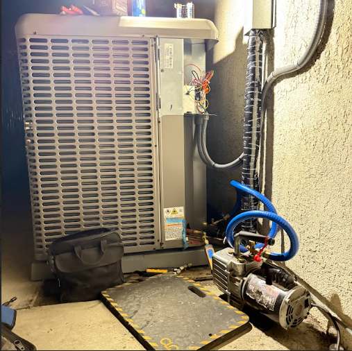 HVAC unit being serviced outdoors; vacuum pump connected with blue hose, tools and bag nearby.