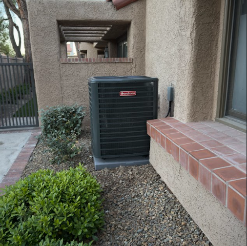 An air conditioning unit next to a building, surrounded by landscaping and gravel.