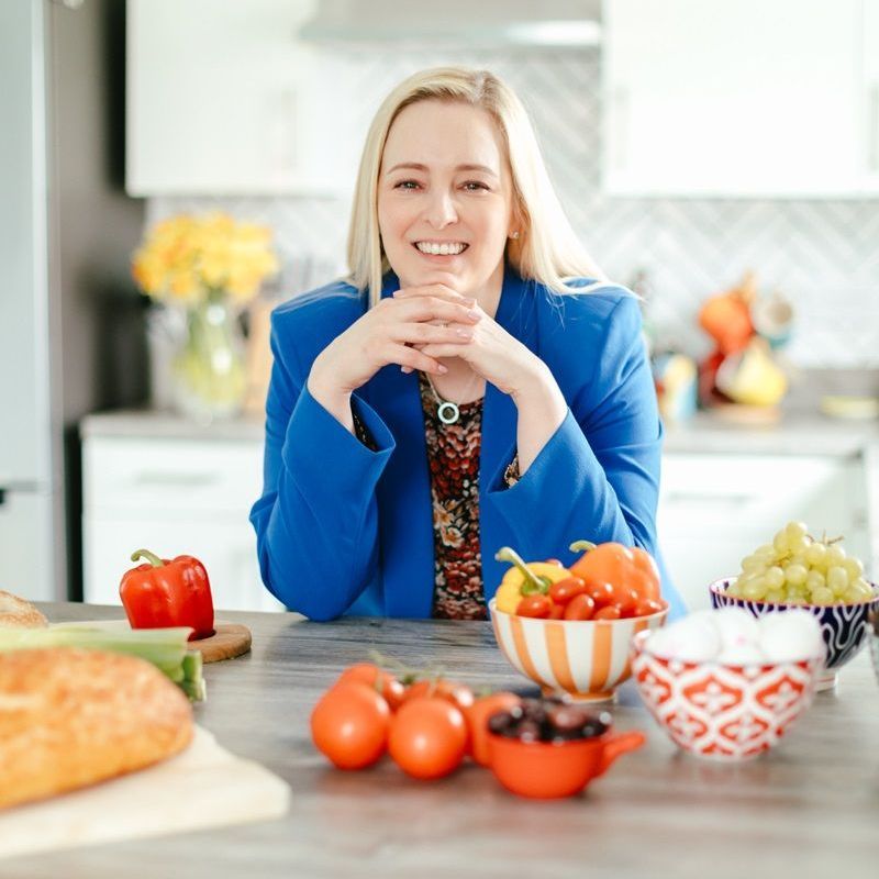 A woman in a blue jacket sits at a table with bowls of fruit and vegetables