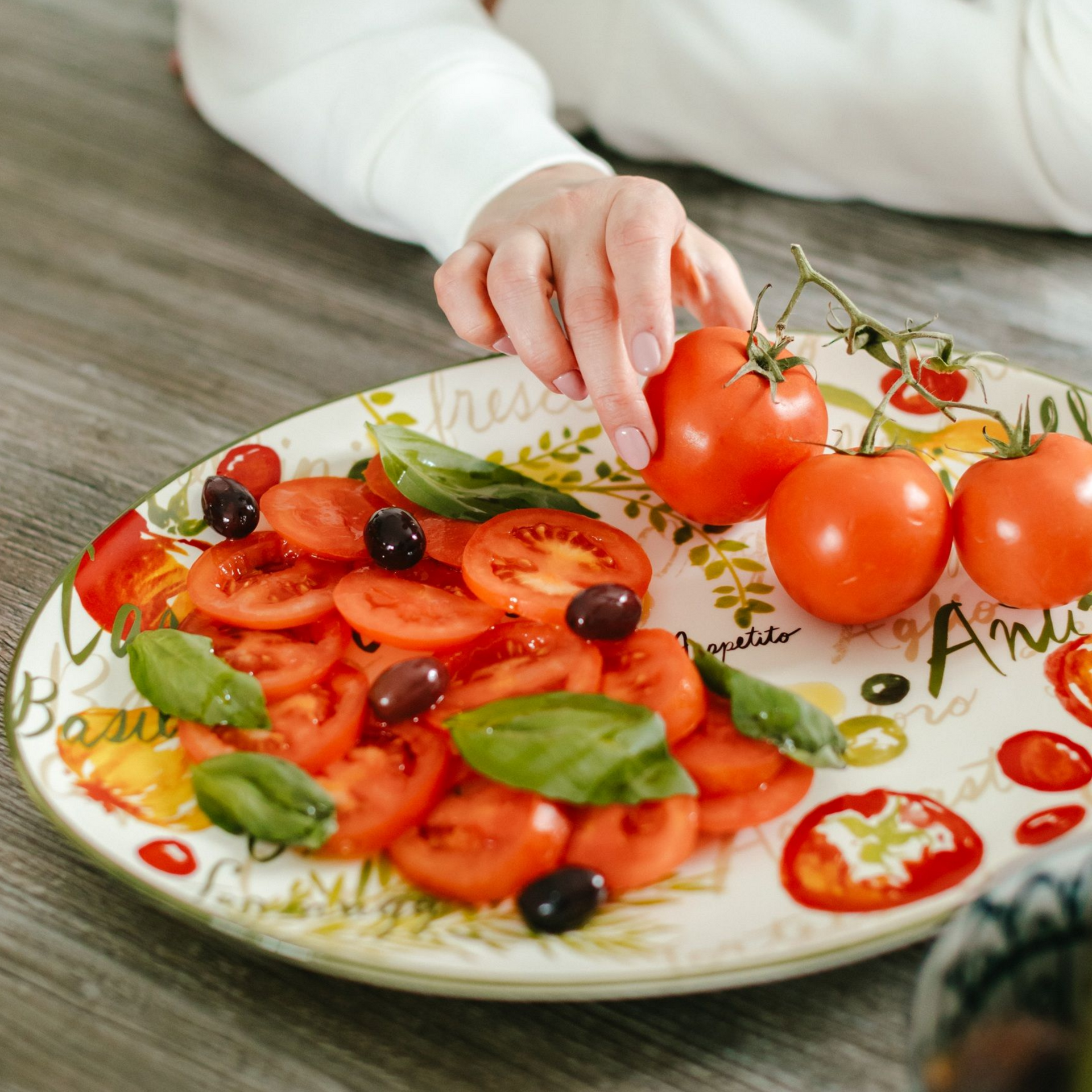 Person arranging sliced tomatoes, basil, and olives on a decorated plate.