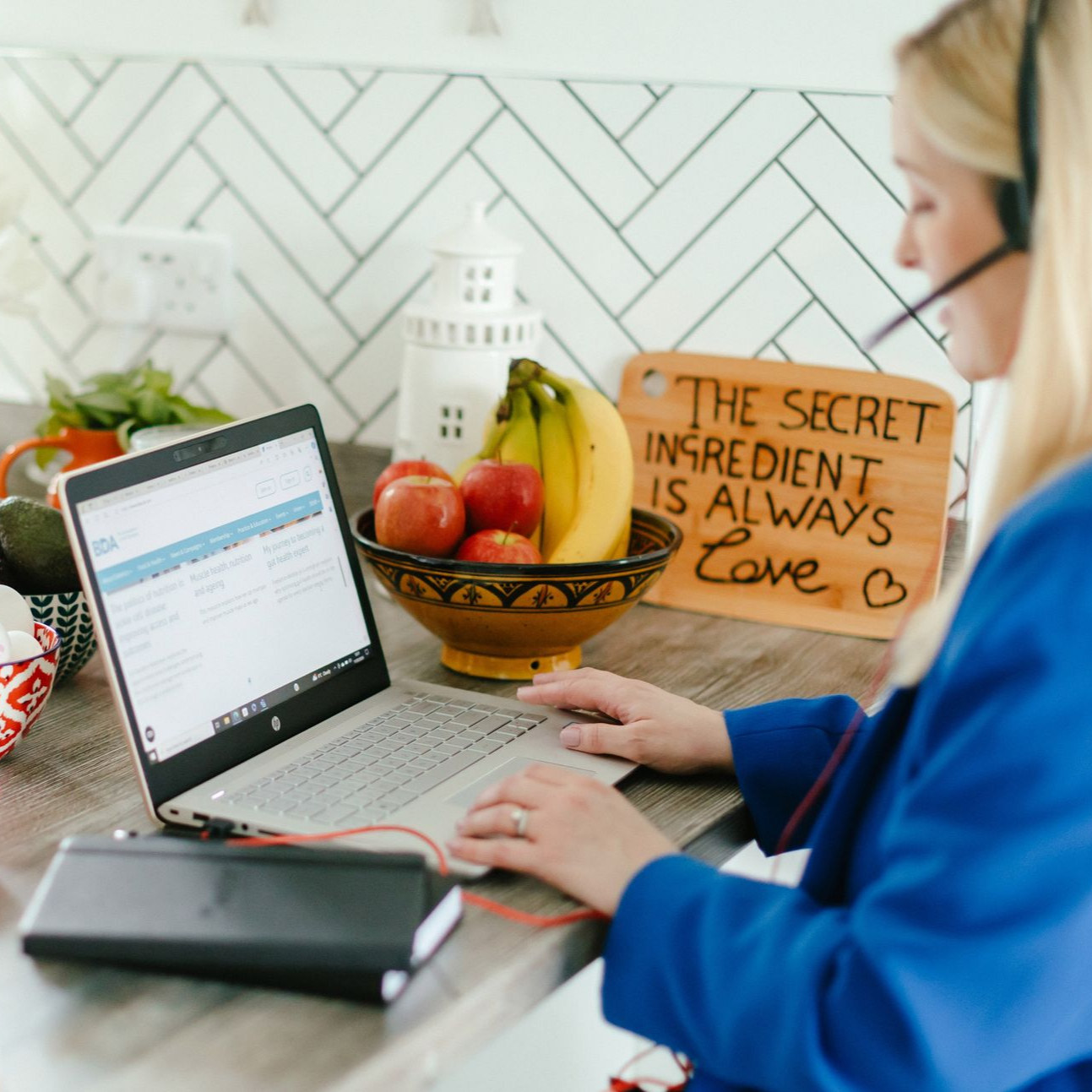Woman working on laptop in kitchen, wearing headset. Fruit bowl, sign