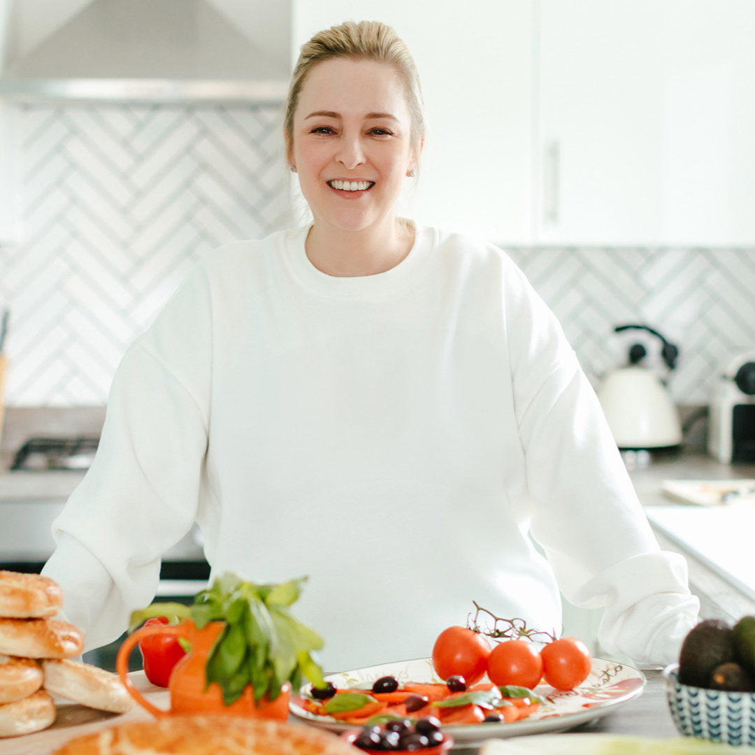 Woman smiling in a kitchen, surrounded by food. Bright setting with a white sweatshirt.