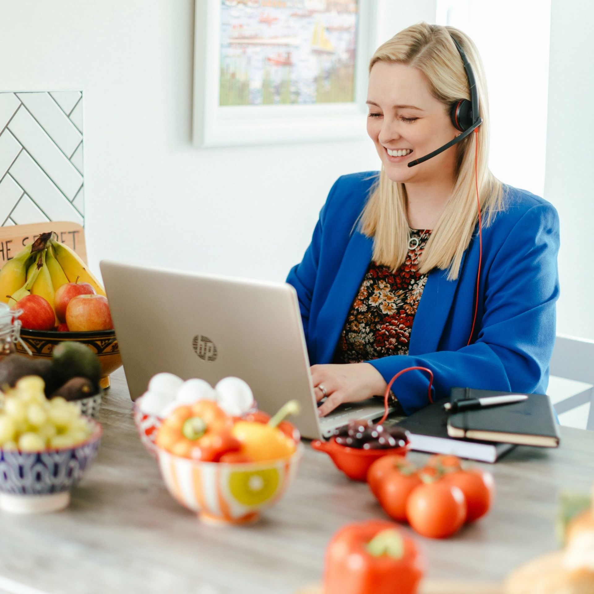 Woman in blue blazer works on laptop with headset; fruit and veggies on table.