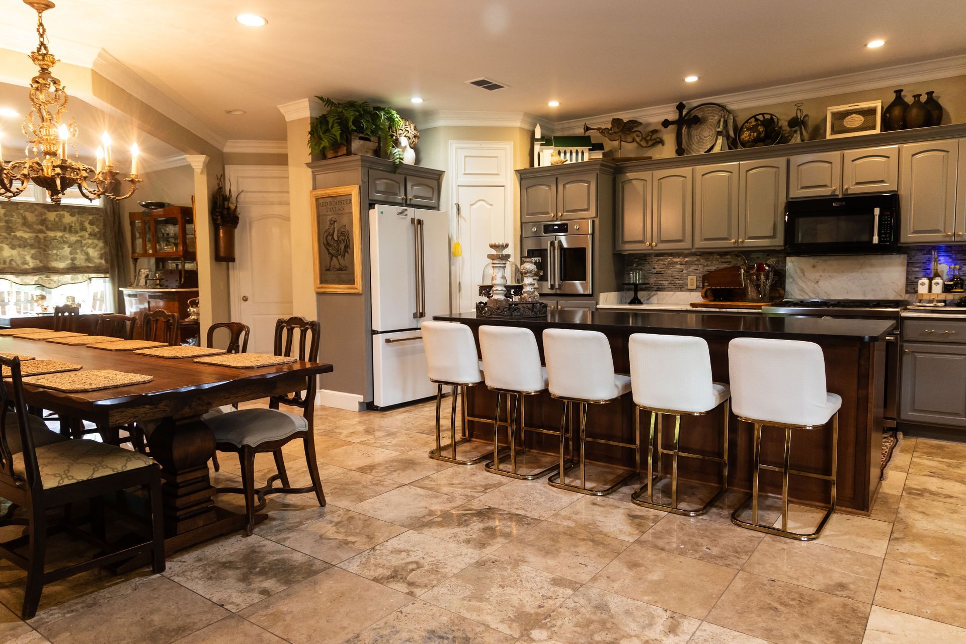 A spacious kitchen with a dark wood island, white bar stools, grey cabinets, and an adjacent dining area with stone floors.