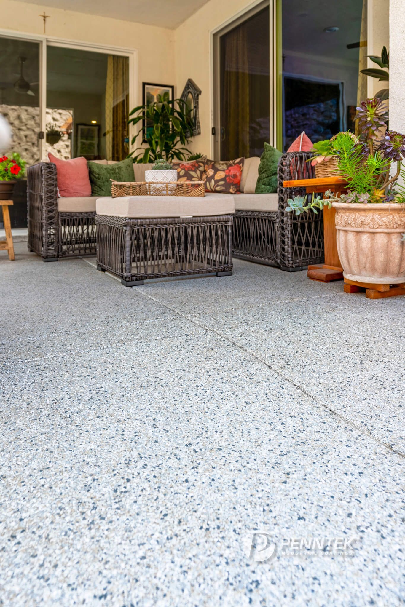 A covered patio features wicker seating with beige cushions, a matching ottoman, and a potted plant on a speckled floor.