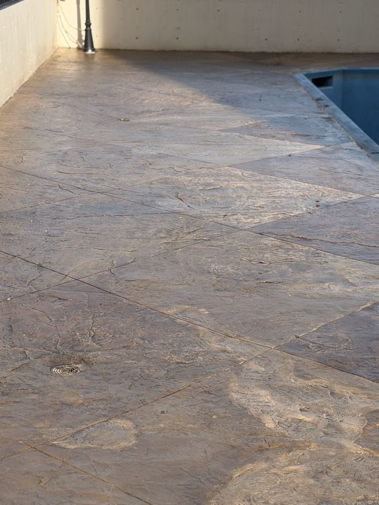Concrete pool deck beside a swimming pool, with a textured stone-like surface and white wall edge