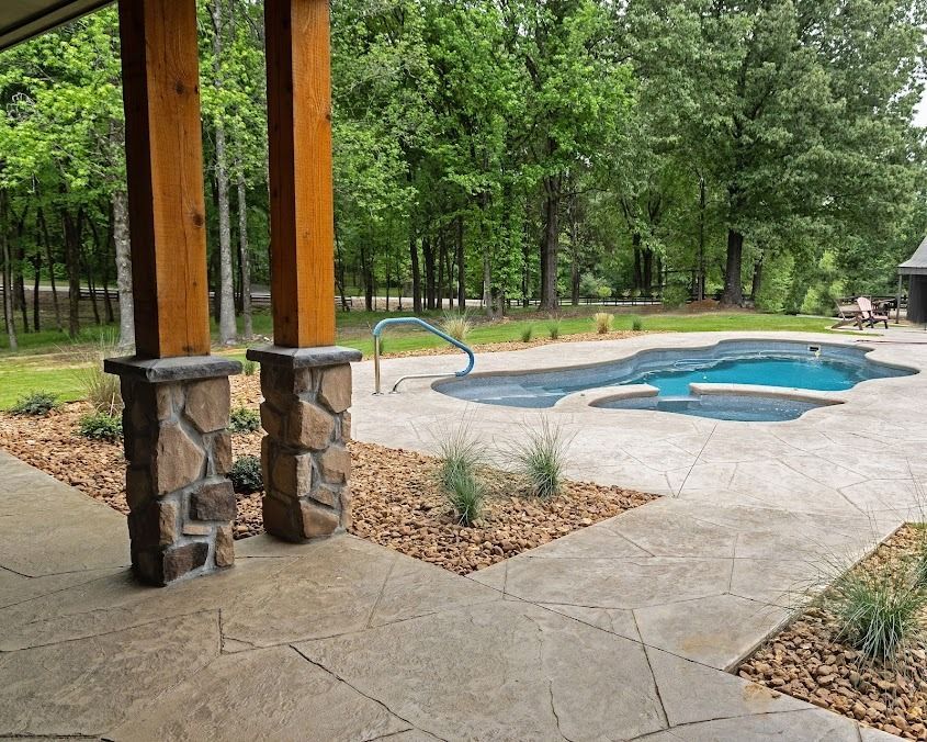 A stone-paved patio with two stone pillars leads to an outdoor swimming pool surrounded by woods.